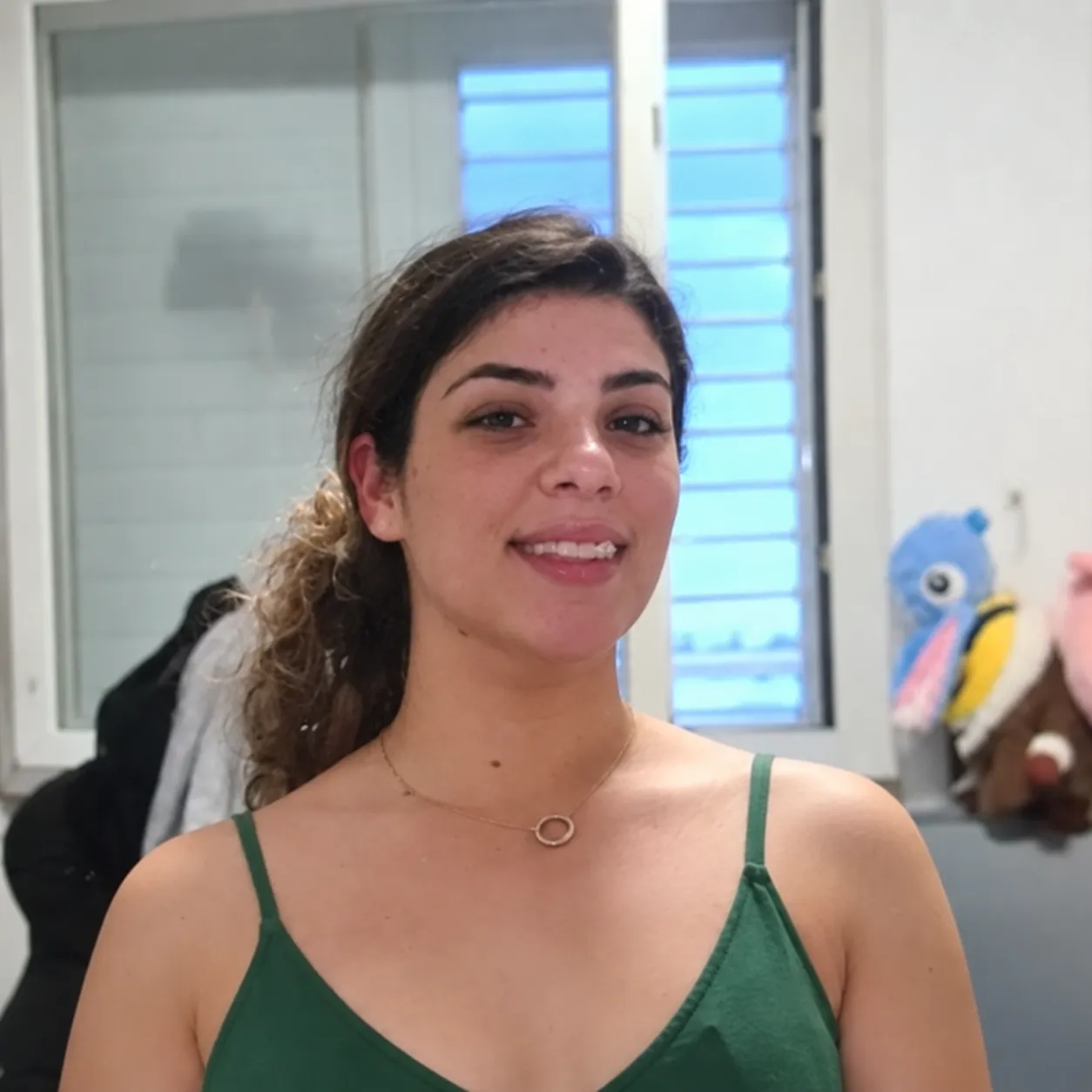 Smiling young woman with curly hair tied back, wearing a green sleeveless top and a delicate necklace, standing indoors near a window with blue light outside and a few plush toys in the background.