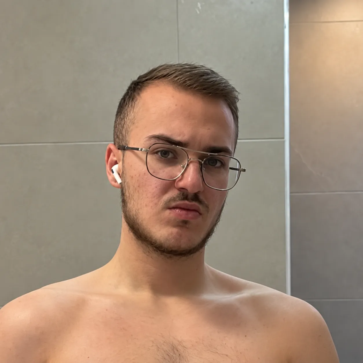 Young man with short light brown hair, a beard, and glasses, wearing white wireless earphones, looking at the camera with a serious expression, standing shirtless against a tiled wall background.