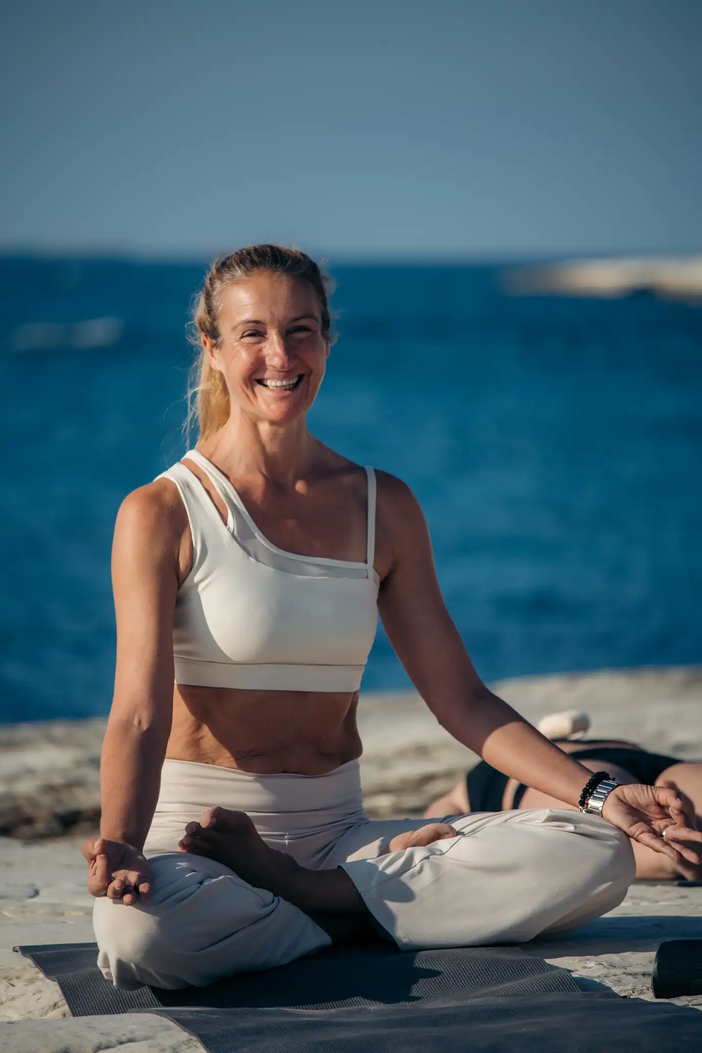 Smiling woman sitting cross-legged on a yoga mat outdoors by the sea, practicing meditation.