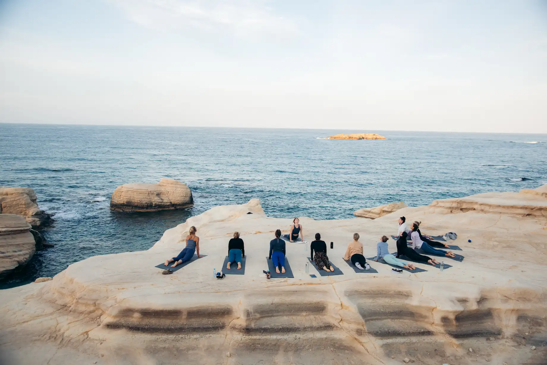 Group yoga session on mats on rocky cliffs overlooking the ocean at sunrise or sunset.