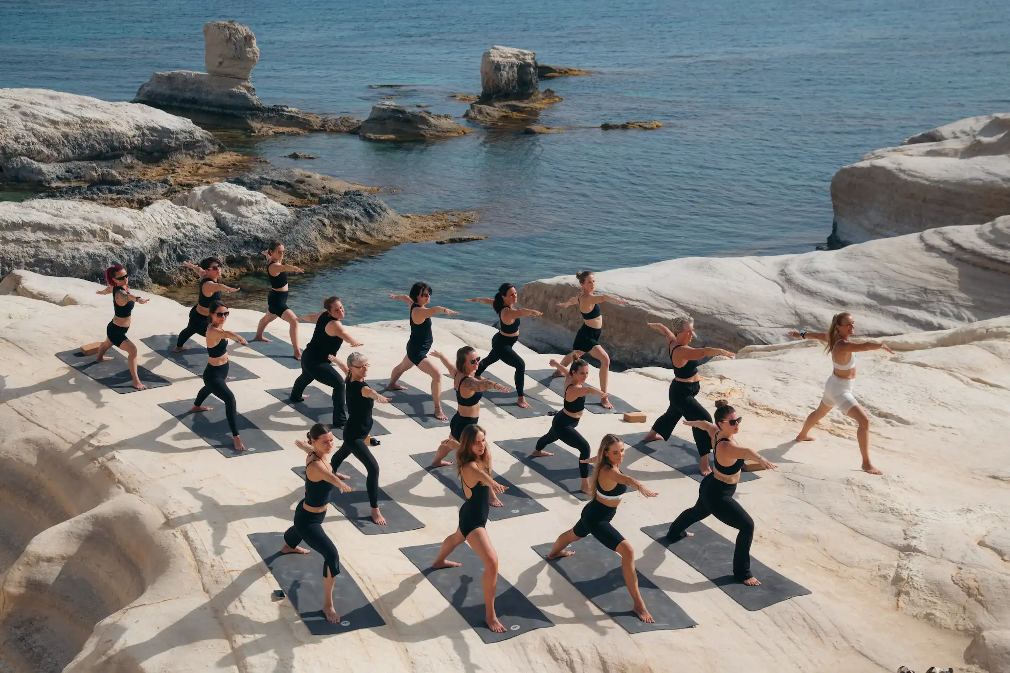 Group of women practicing yoga in warrior pose on mats atop white rocky cliffs by the sea.