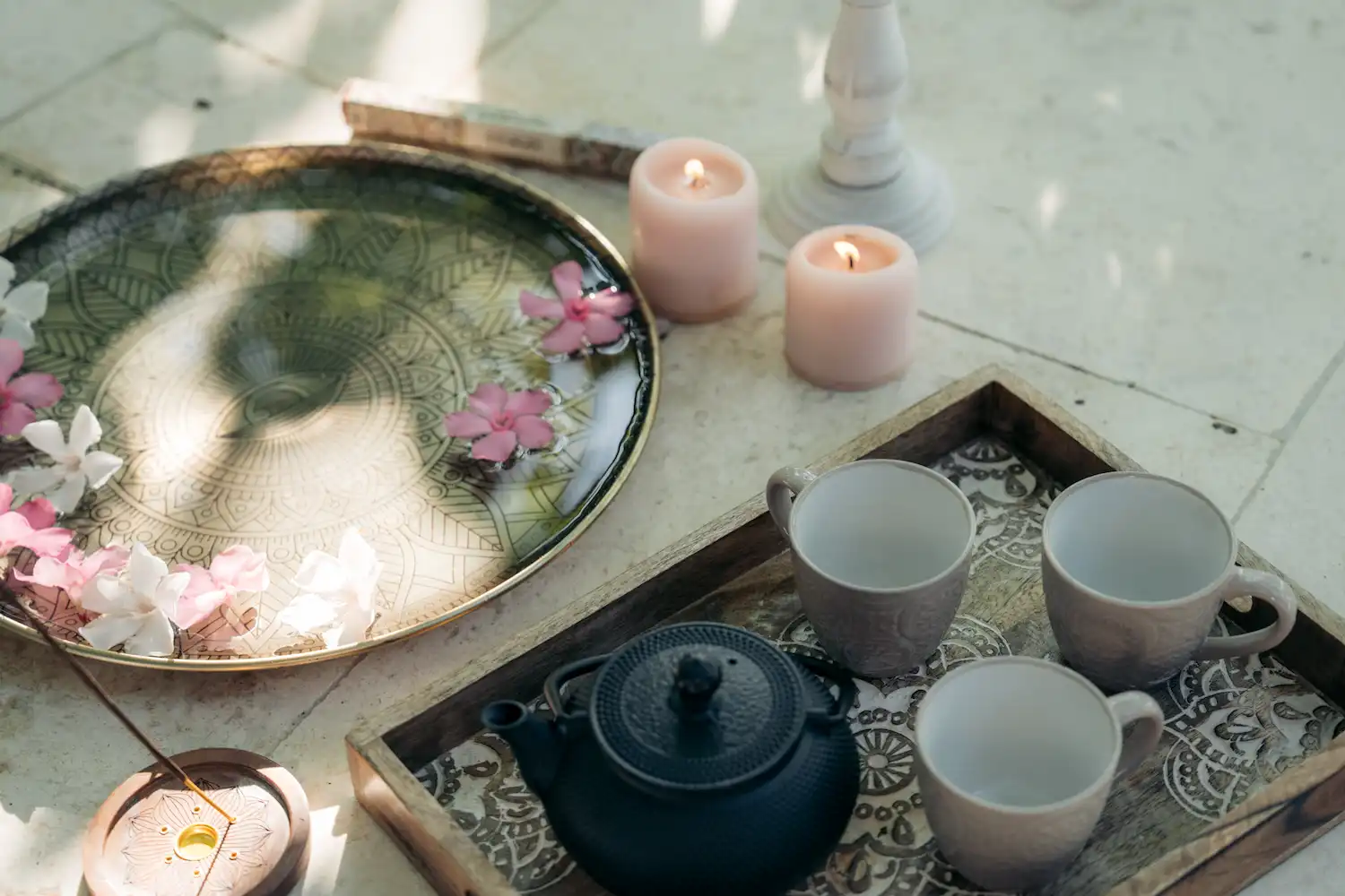 Black teapot and three empty cups on a patterned wooden tray beside a decorative plate with floating pink and white flowers and two lit candles.