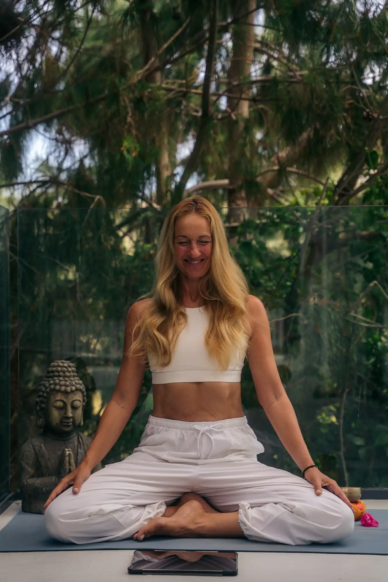 Smiling woman with long blonde hair sitting cross-legged on a yoga mat outdoors with a Buddha statue and greenery in the background.
