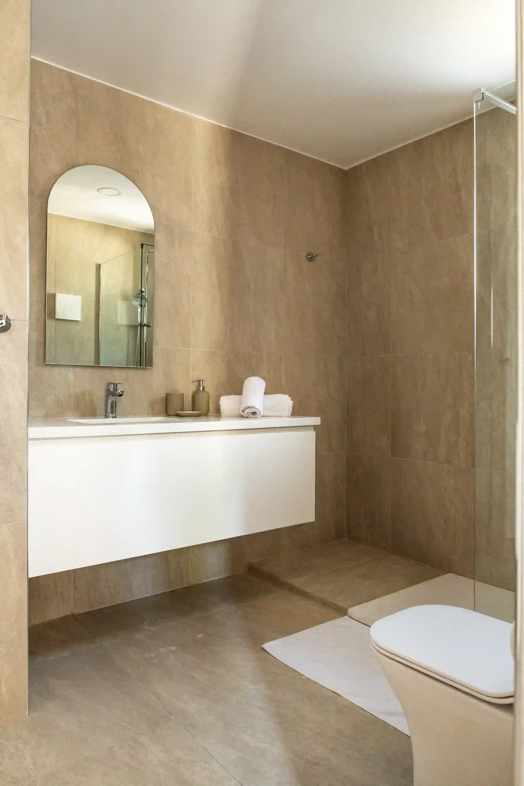 Minimalist bathroom with beige tiles, a white wall-mounted vanity with a round-top mirror, rolled white towels, and a walk-in shower.