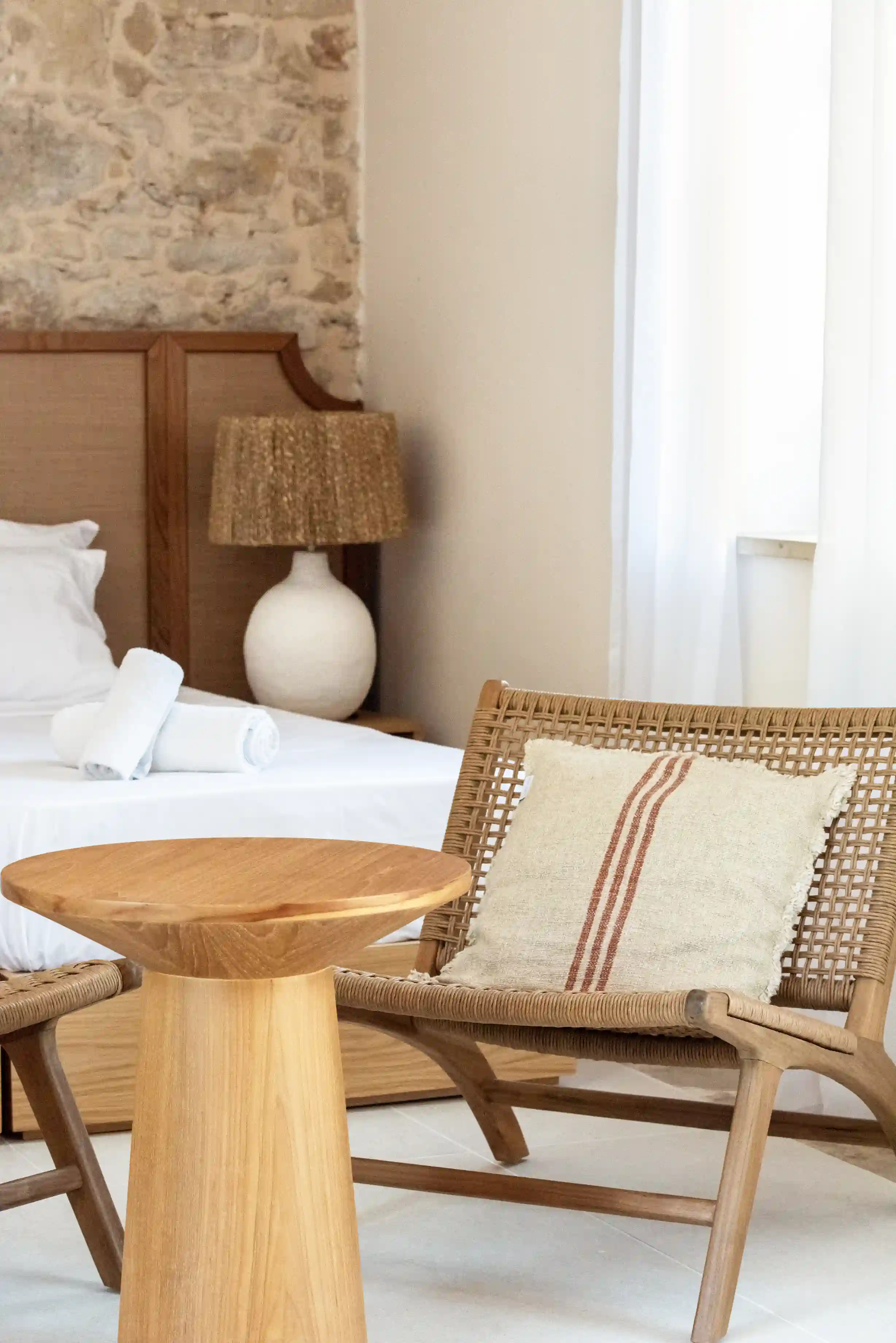 Minimalist bedroom corner with wicker chair featuring a beige striped cushion, wooden side table, and bed with white linens and rolled towels.
