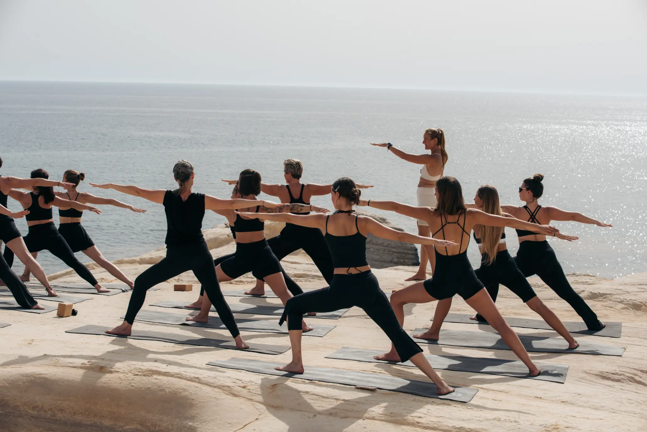 Teilnehmerinnen der Hybrid-Ausbildung strecken die Arme bei Abendlicht auf der Yoga Shala auf Zypern, die Lehrerin leitet die Praxis an