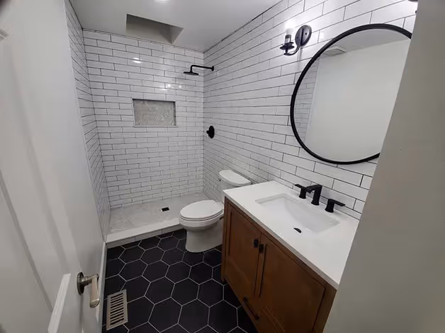 Modern bathroom with white subway tile walls, black hexagonal floor tiles, a walk-in shower with black fixtures, a wooden vanity with white countertop, and a round mirror.
