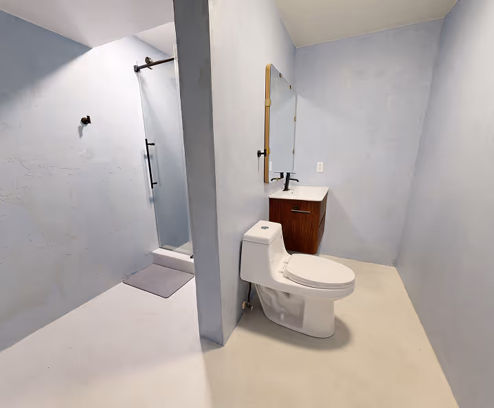 Minimalist bathroom with light gray walls, a white toilet, a wooden vanity with a black faucet, and a walk-in shower with glass door.