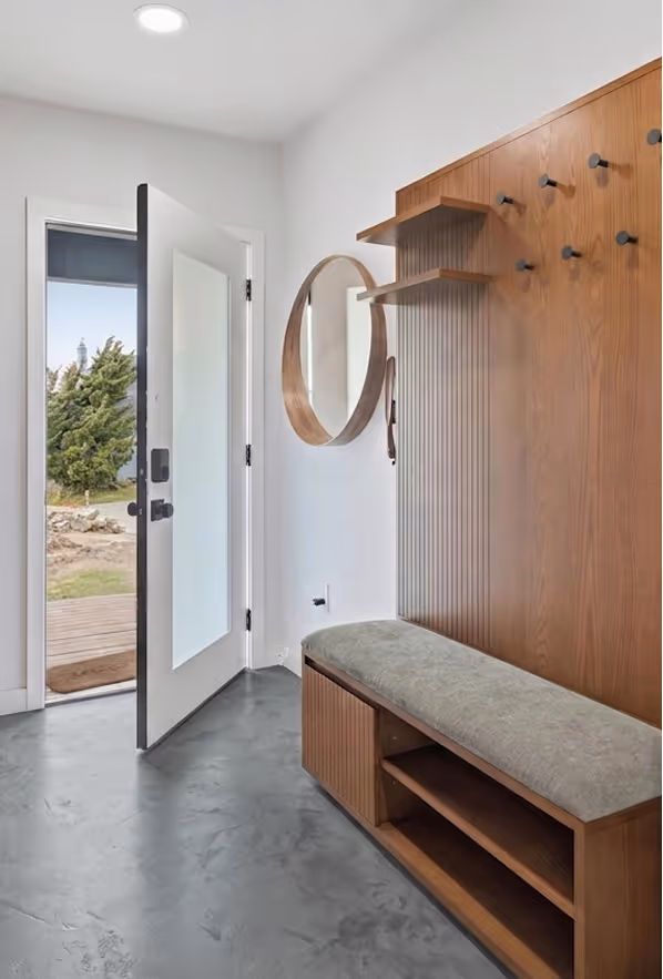 Entryway with open white door, concrete flooring, and wood bench with cushioned seat and coat hooks.