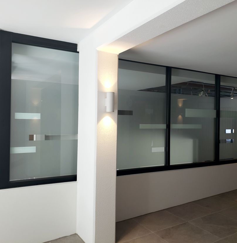 Interior view of a modern office corridor with frosted glass windows featuring horizontal clear stripes and a wall-mounted light fixture casting upward and downward light on a white column.
