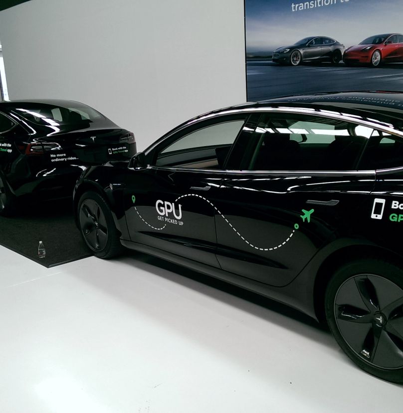 Two black Tesla electric cars parked indoors on a white floor, with one car displaying 'GPU GET PICKED UP' and a graphic of a dotted route ending in an airplane icon, promoting a ride service app.
