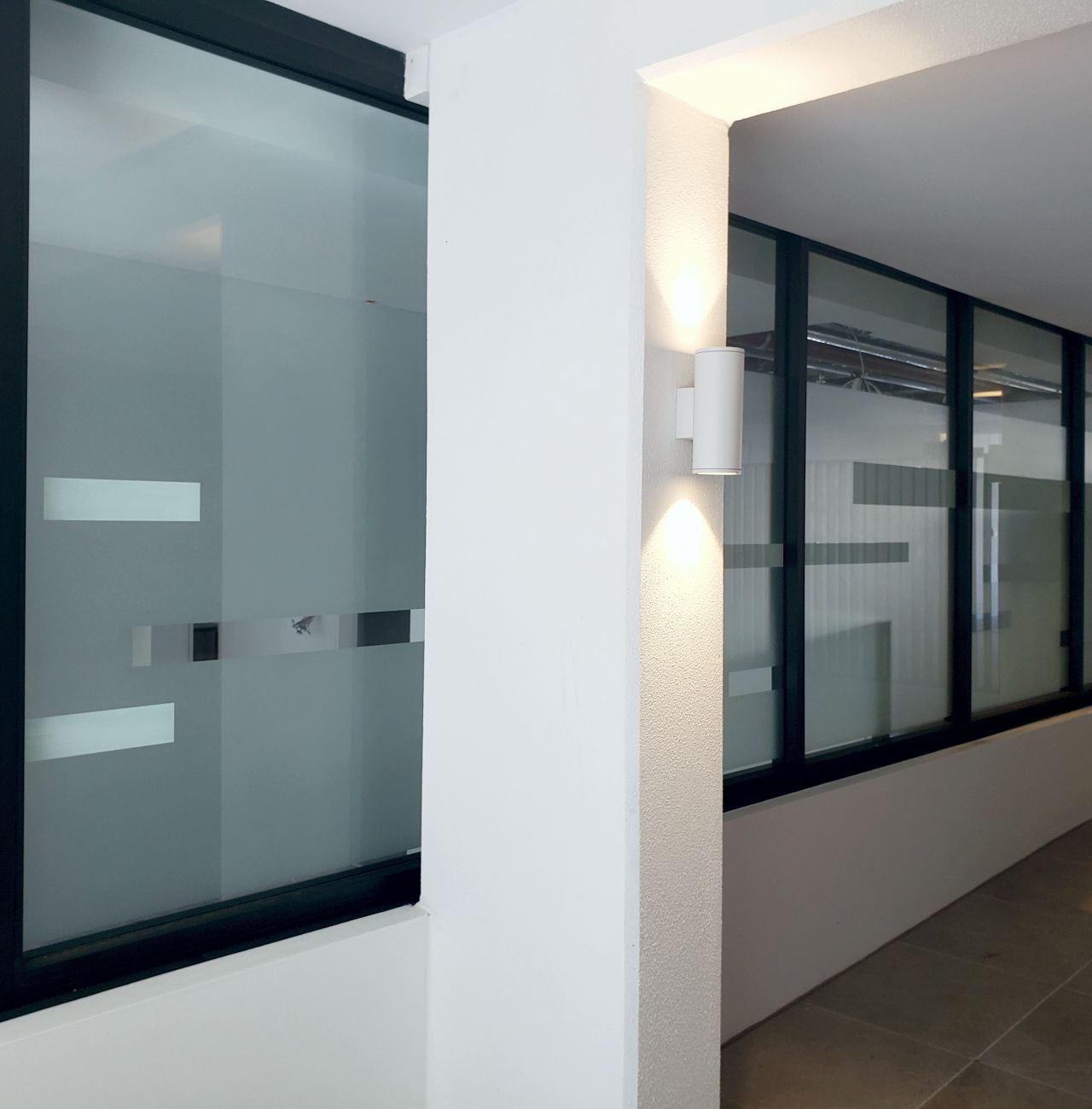 Indoor corridor with large frosted glass windows framed in black and a white wall with a mounted cylindrical light fixture casting upward and downward light.