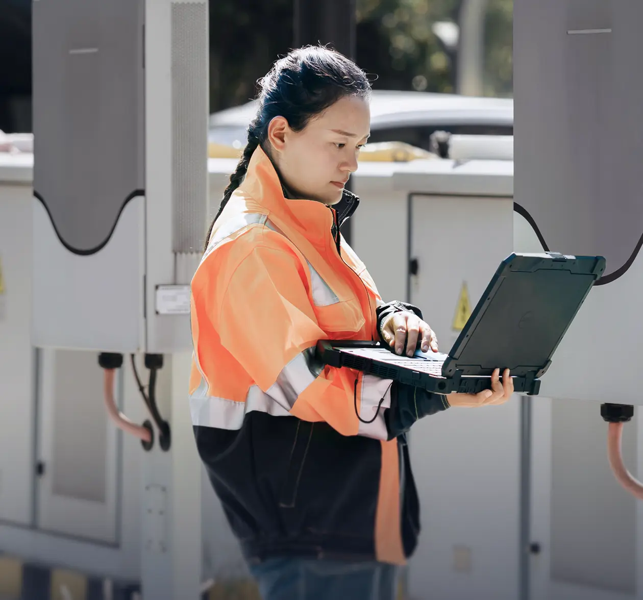 Technician in a high-visibility jacket using a laptop outdoors near electrical equipment.