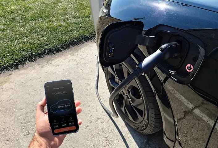Hand holding a smartphone displaying electric vehicle charging status next to a black car plugged into a charging port.
