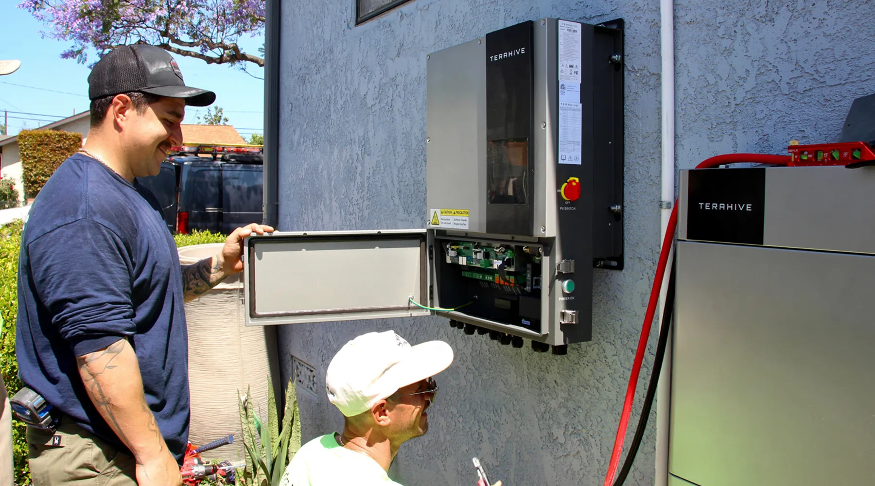 Two men inspecting an open electrical panel mounted on a house exterior wall.