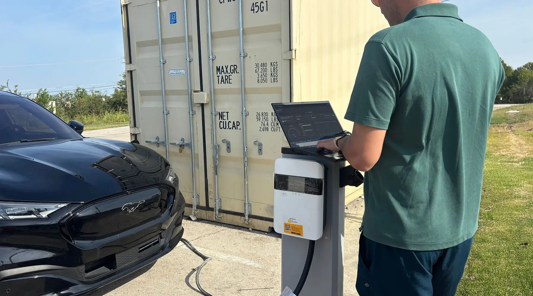 Man in green shirt using a laptop on a charging station next to a E car outdoors.