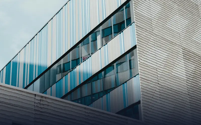 A modern building facade with clean geometric lines, featuring reflective glass windows and metal panels under soft natural light.