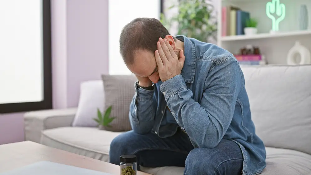Mann sitzt mit gesenktem Kopf und hält sich den Kopf vor Schmerzen – Symbolbild für Cannabis Überdosis