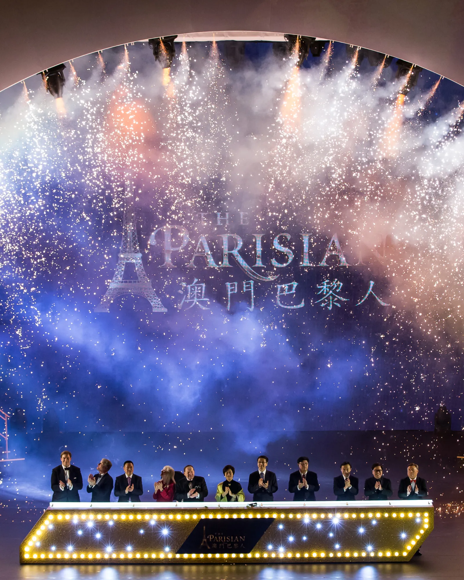 Performer in glittery jacket and colorful pants with large wing-like props on stage, with illuminated cityscape backdrop.