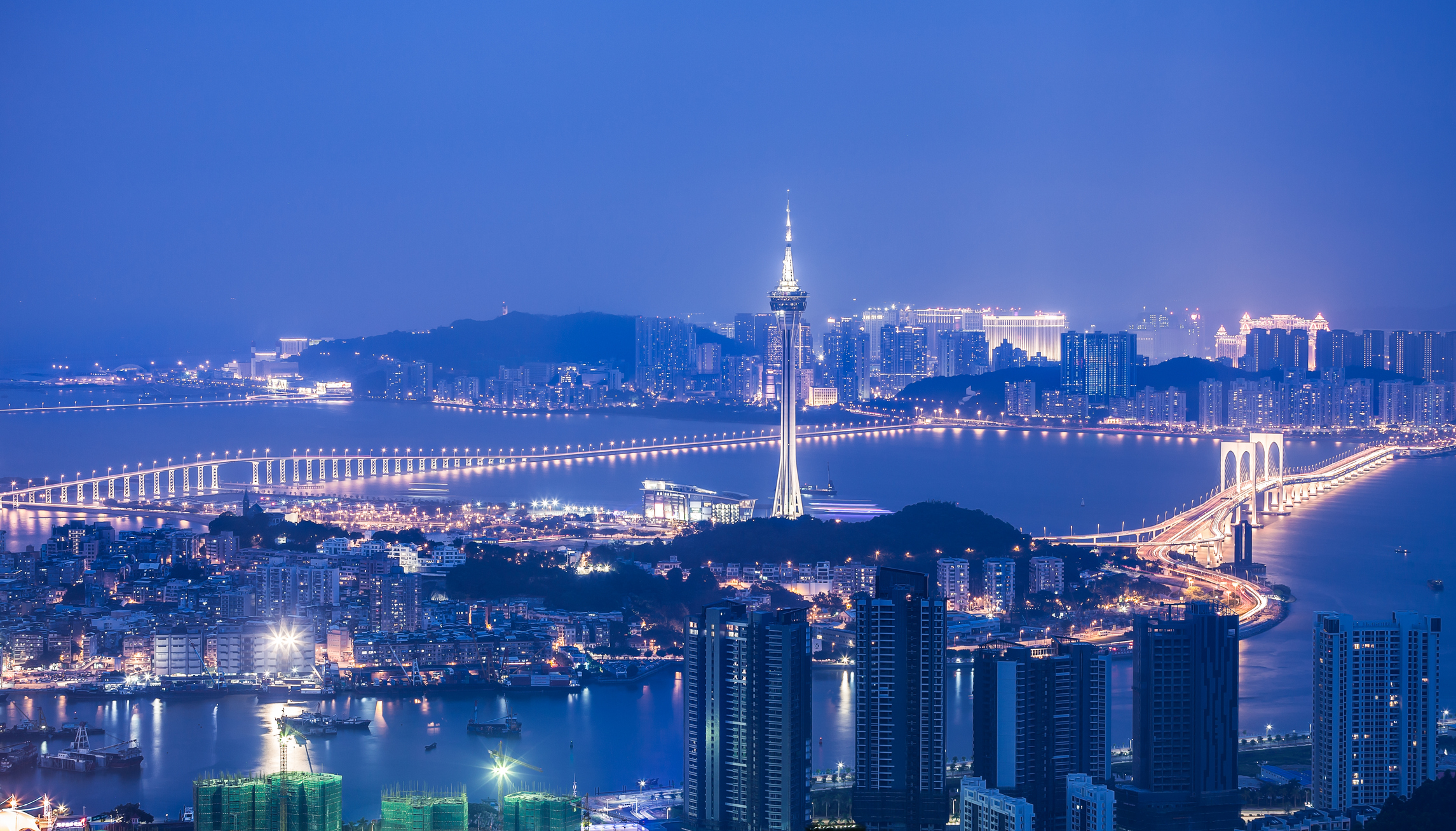 Night view of a city skyline with a tall illuminated tower and a lit bridge stretching across water.