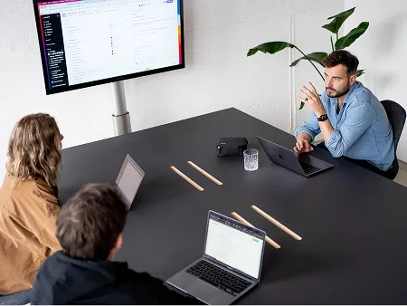 Meeting in einem modernen Konferenzraum: Drei Teammitglieder sitzen mit Laptops um einen Tisch und diskutieren mit einem Kollegen, der ihnen gegenüber sitzt. Im Hintergrund zeigt ein großer Bildschirm digitale Arbeitsinhalte. Eine große Zimmerpflanze sorgt für eine angenehme Atmosphäre.