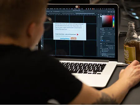 Ein Mitarbeiter arbeitet am Laptop an einer Designsoftware für eine Kampagne, neben ihm steht eine Club Mate Flasche auf dem Schreibtisch – kreativer Arbeitsalltag in der Agentur.