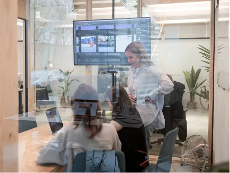 Blick durch eine Glasscheibe in einen modernen Besprechungsraum: Zwei Personen sitzen an einem Holztisch mit Laptops und arbeiten gemeinsam an einem Projekt. Eine Frau steht neben dem Tisch und erklärt etwas, während auf einem großen Monitor an der Wand verschiedene digitale Kampagnen und Layouts angezeigt werden. Im Hintergrund sind Pflanzen und weitere Arbeitsplätze sichtbar. Die Szene vermittelt Teamarbeit und kreative Zusammenarbeit in einer Agentur.