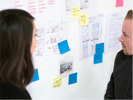Zwei Teammitglieder besprechen Wireframes und Konzeptskizzen an einer Whiteboard-Wand mit Post-its – kollaborative Strategie- und Konzeptentwicklung in der Agentur.