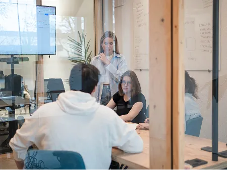 Drei Personen sitzen in einem modernen Meetingraum an einem Holztisch und besprechen gemeinsam Notizen und Strategien, während eine weitere Frau stehend zuhört und nachdenkt. Durch die Glasscheibe sind Whiteboards mit Stichpunkten und ein Bildschirm sichtbar. Die Szene zeigt konzentrierte Teamarbeit und Strategieentwicklung in einer Agentur.