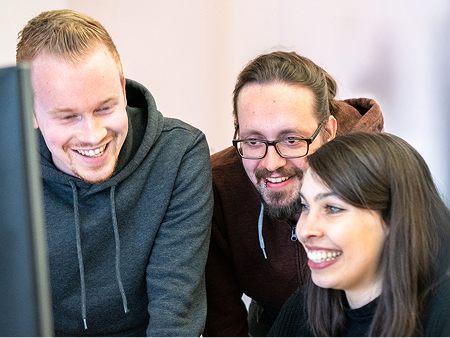 Drei Kolleg:innen sitzen gemeinsam vor einem Bildschirm, lachen und schauen konzentriert auf den Monitor. Die entspannte und freundliche Atmosphäre unterstreicht das Teamwork und die Freude an der Zusammenarbeit innerhalb der Agentur.