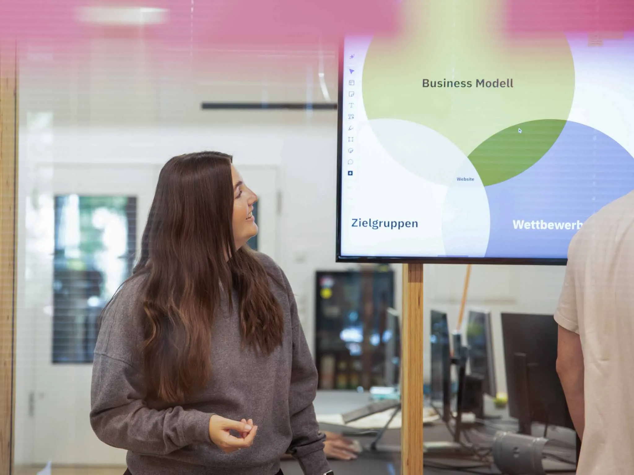 Eine Frau präsentiert in einem Büro eine Folie von einem großen Screen mit überlappenden Kreisen zu Business-Modell, Zielgruppen und Wettbewerb.