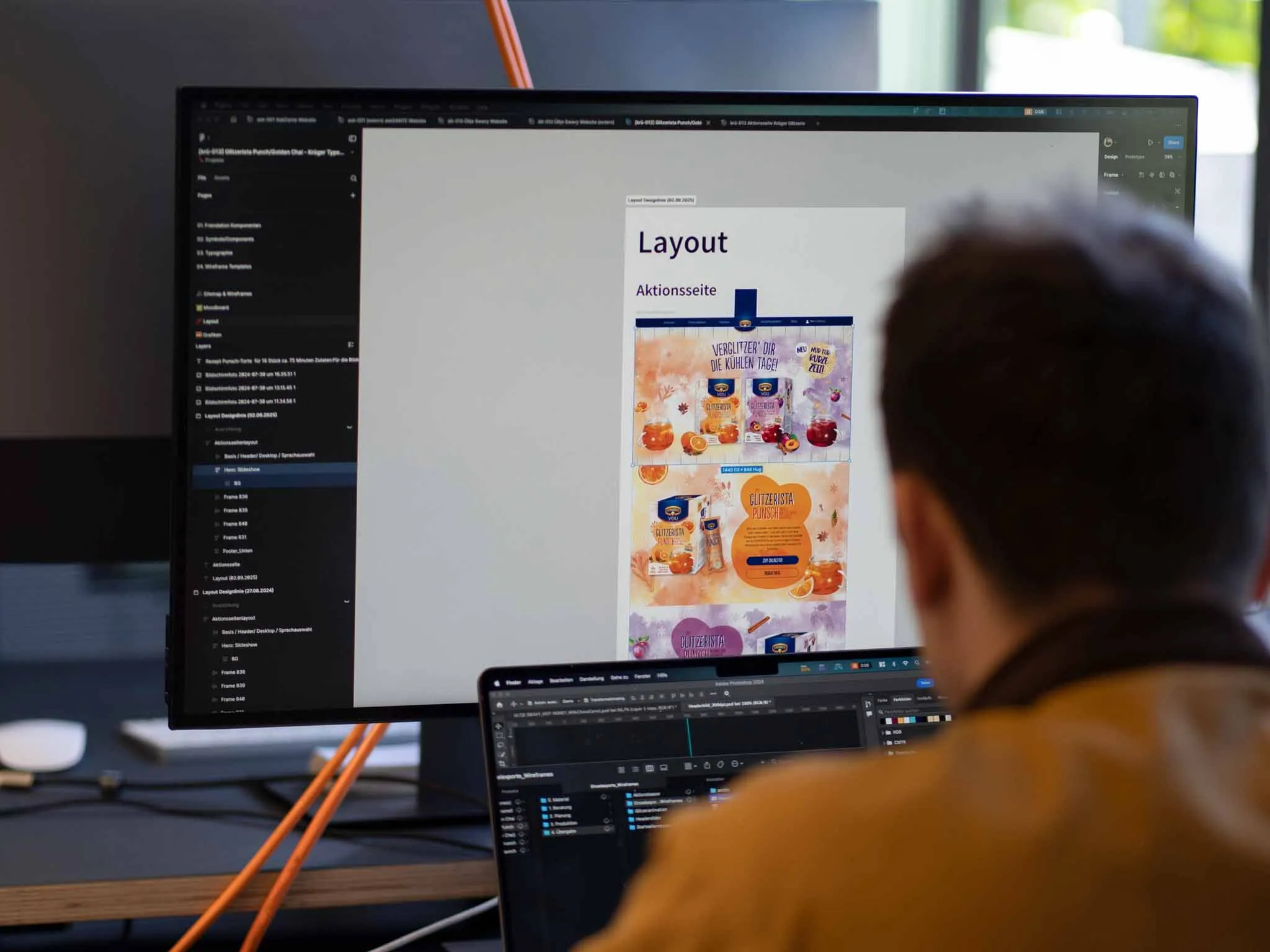 Ein Designer sitzt an einem Schreibtisch vor zwei Bildschirmen, auf denen digitale Assets und Layout-Kreationen zu sehen sind.