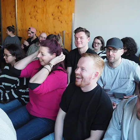 Das Bild zeigt ausgelassene Stimmung auf einer Versammlung im Office. Im Hintergrund sieht man Performance Marketing Manager Meo in die Kamera lächeln, während Projektmanager Stephan im Vordergrund ausgelassen lacht.