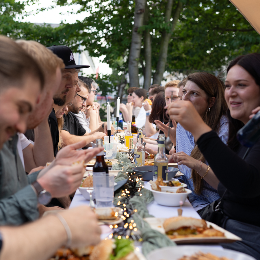  Eine Momentaufnahme des Adfriends Sommerfests. Die Kolleg:innen sitzen an einer großen Tafel, die gedeckt mit verganen Köstlichkeiten ist und heben das Glas um gemeinsam anzustoßen.