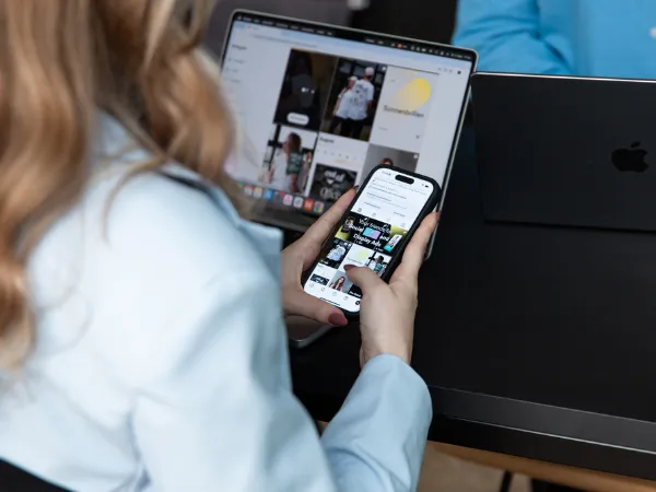 Frau mit  einem IPhone in der Hand scrollt durch Social-Media-Inhalte, im Hintergrund steht ein Laptop mit geöffnetem Feed auf einem Schreibtisch.