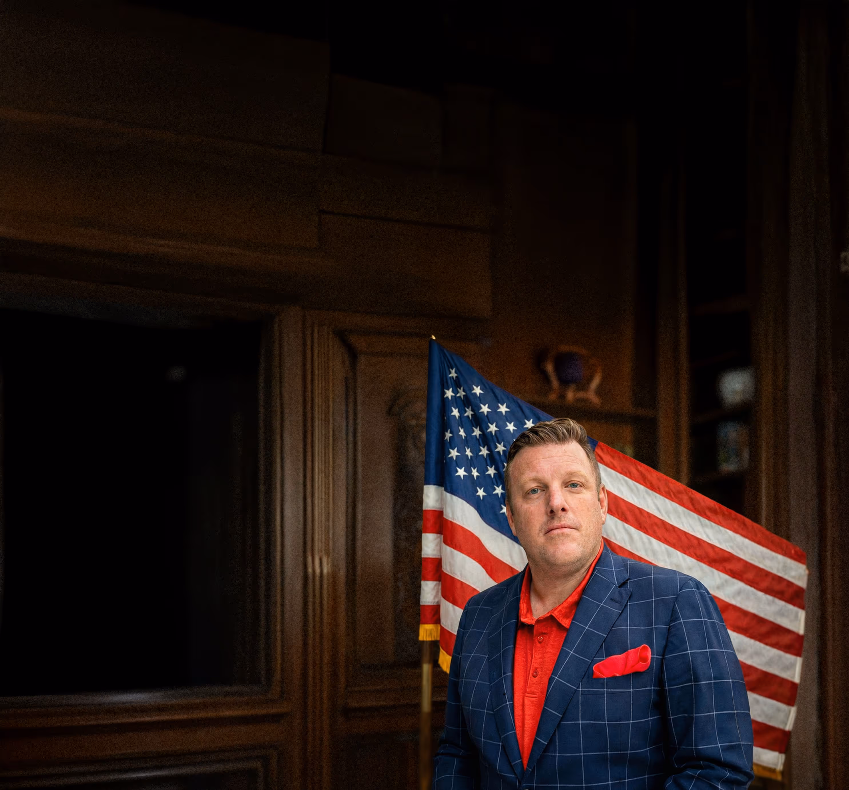 Paul Bondar in a blue checkered suit with a red shirt and pocket square standing in front of an American flag indoors.