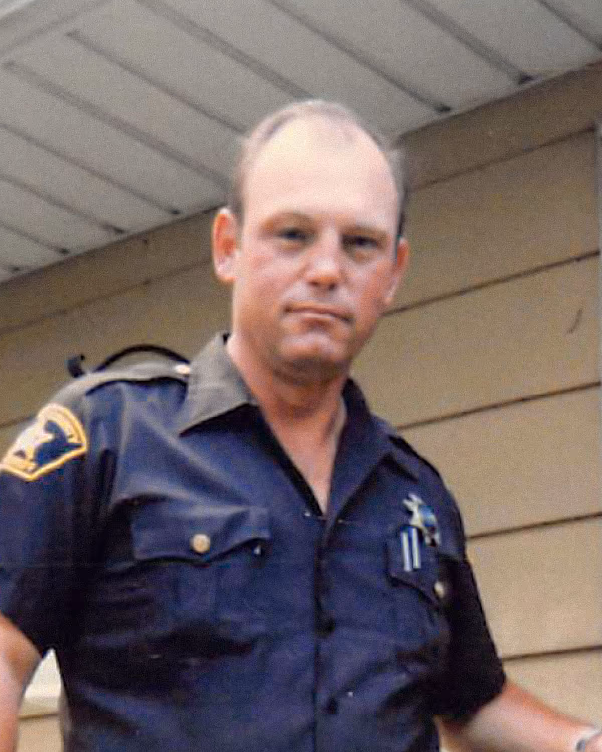 Paul Bondar's dad in a dark police uniform with a badge and shoulder patch standing in front of a beige wooden wall.