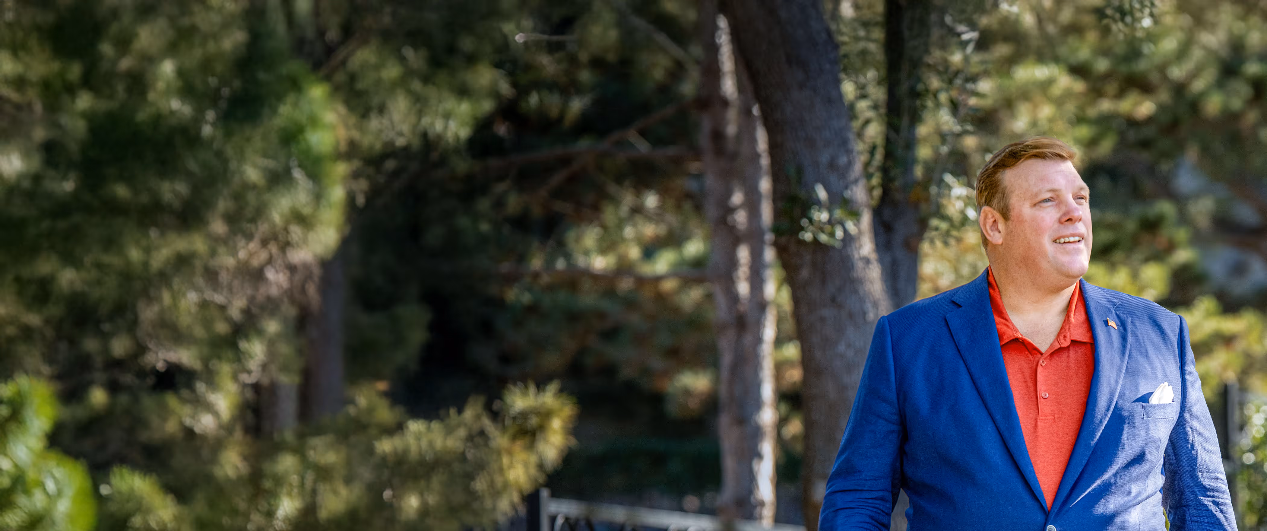 Paul Bondar in blue blazer and red shirt standing outdoors with trees in the background, looking to the right.
