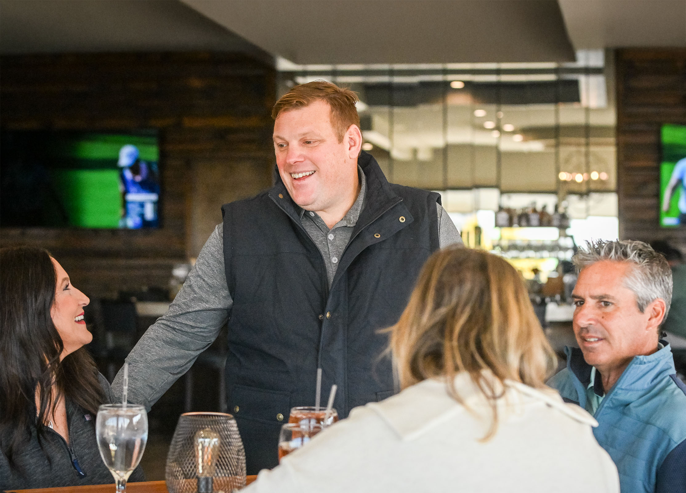 Paul Bondar and a group of three people socializing at a restaurant table with drinks, one man standing and smiling while talking to seated friends.