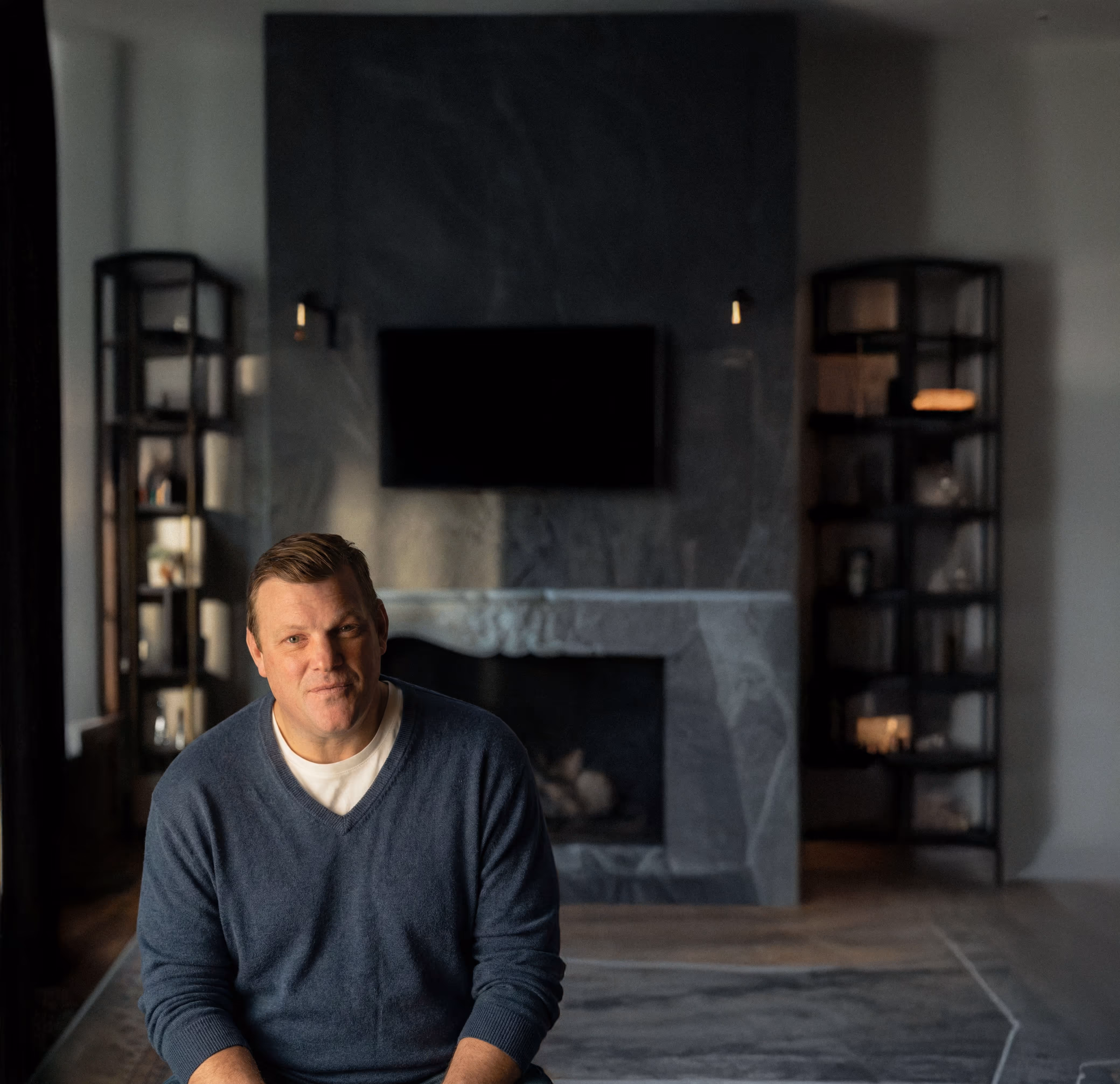 Paul Bondar in a blue sweater sitting indoors in front of a gray stone fireplace with a wall-mounted TV and shelves on each side.