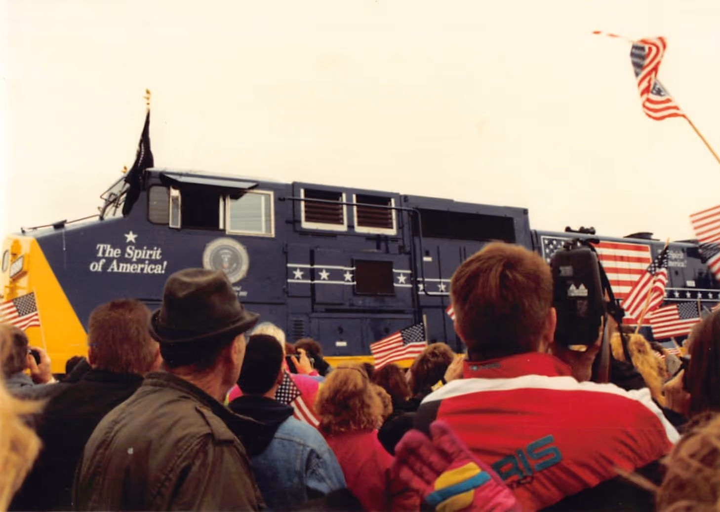 Crowd gathered around a blue and yellow train decorated with American flags and the words 'The Spirit of America!' on its side.