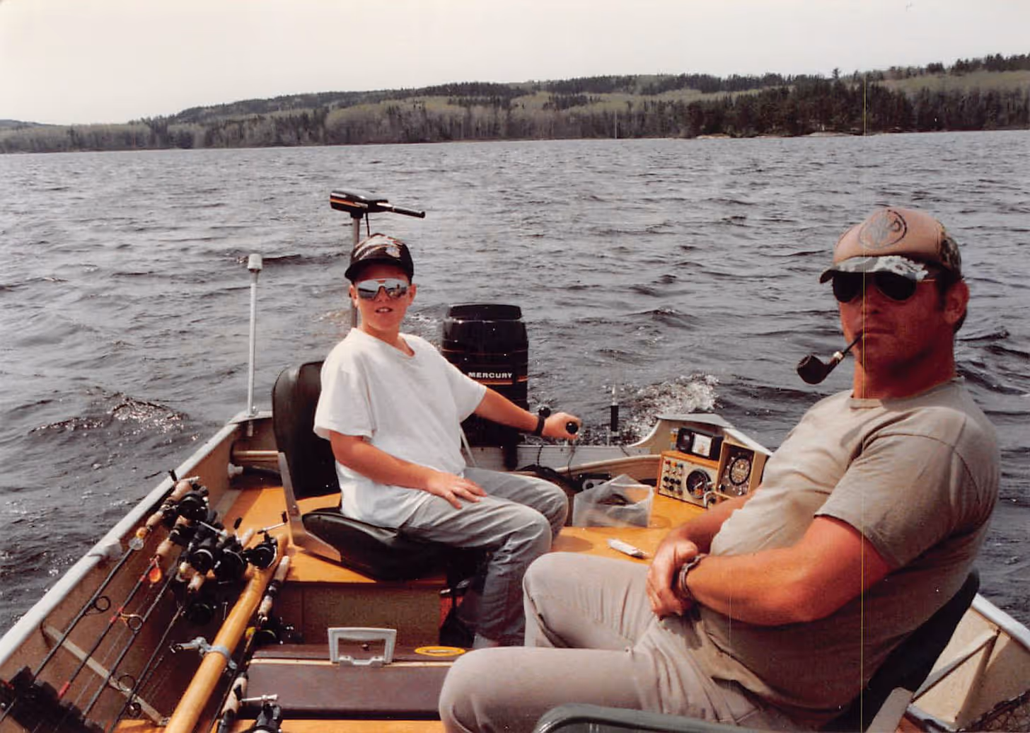 Paul Bondar and his dad on a fishing boat, one steering and the other smoking a pipe, with fishing rods and a lake with forested hills in the background.