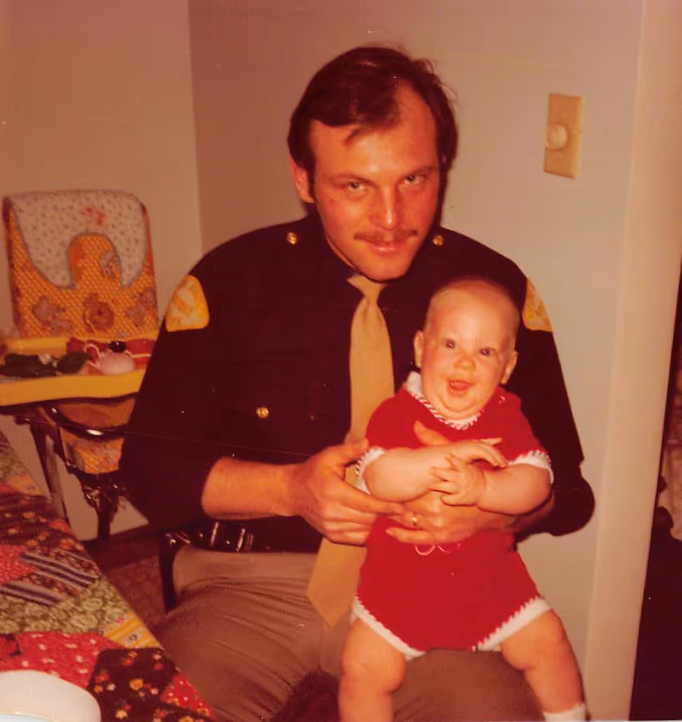 Paul Bondar's dad in police uniform holding a smiling baby dressed in red inside a home.