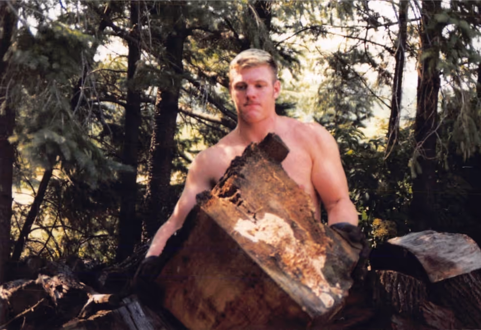 Paul Bondar carrying a large piece of wood in a forested area.