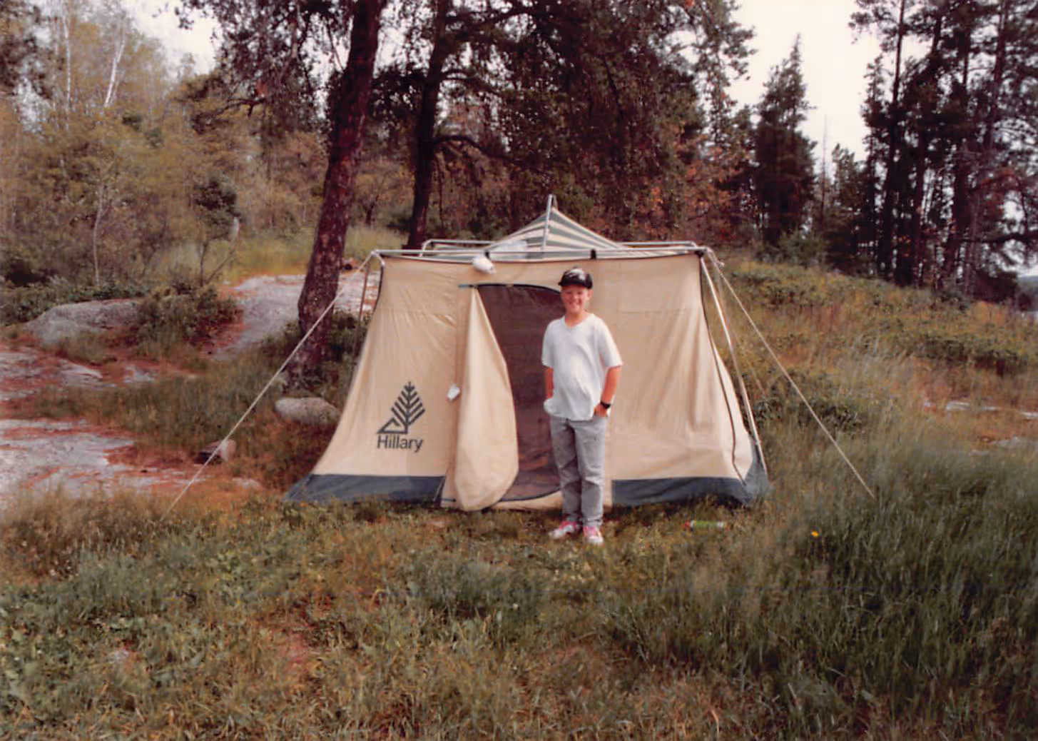 Young boy Paul Bondar wearing a white t-shirt and gray pants stands smiling in front of a beige Hillary tent in a grassy wooded camping area.