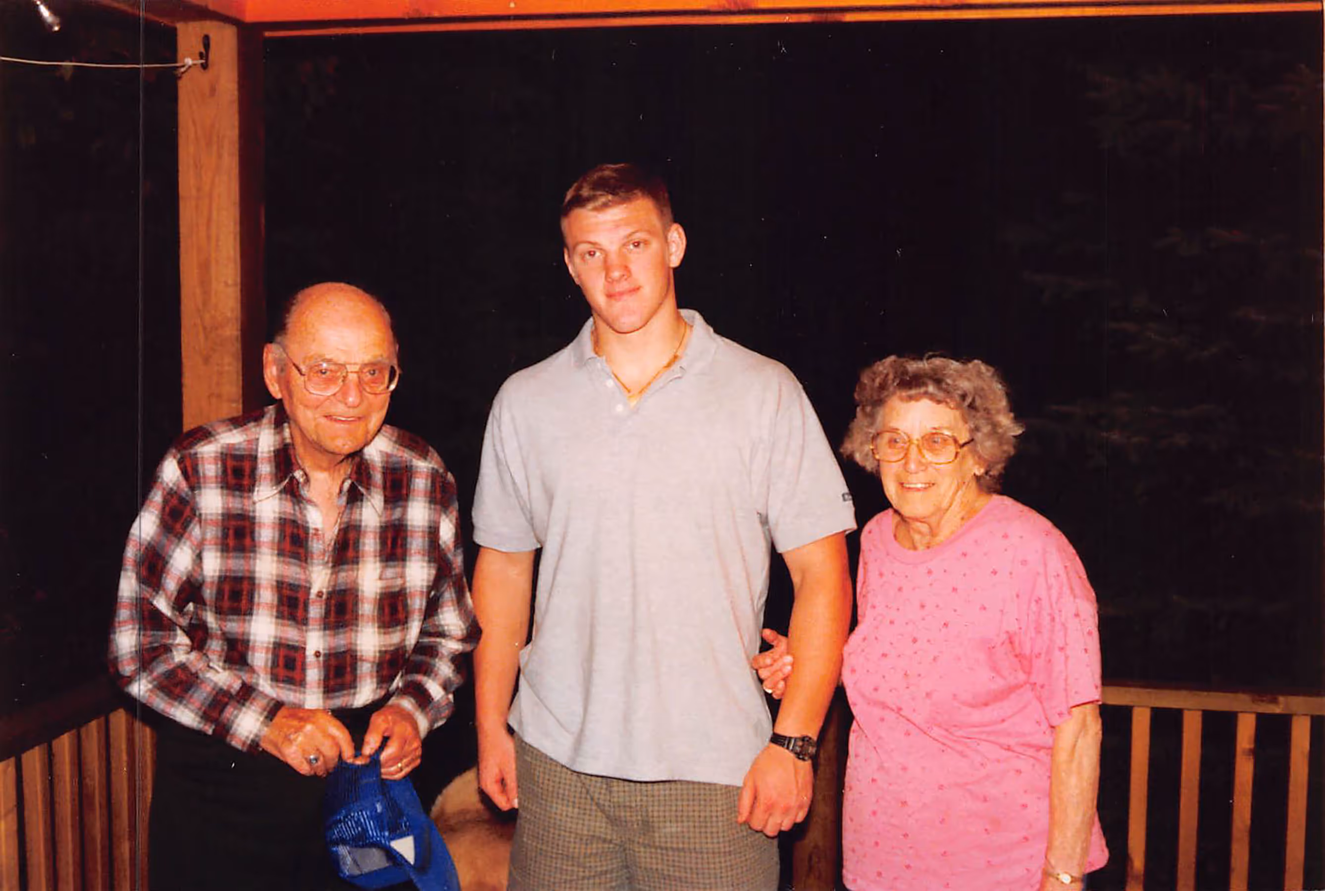 Young man Paul Bondar standing between an elderly man holding a blue cap and an elderly woman in a pink shirt on a wooden porch at night.