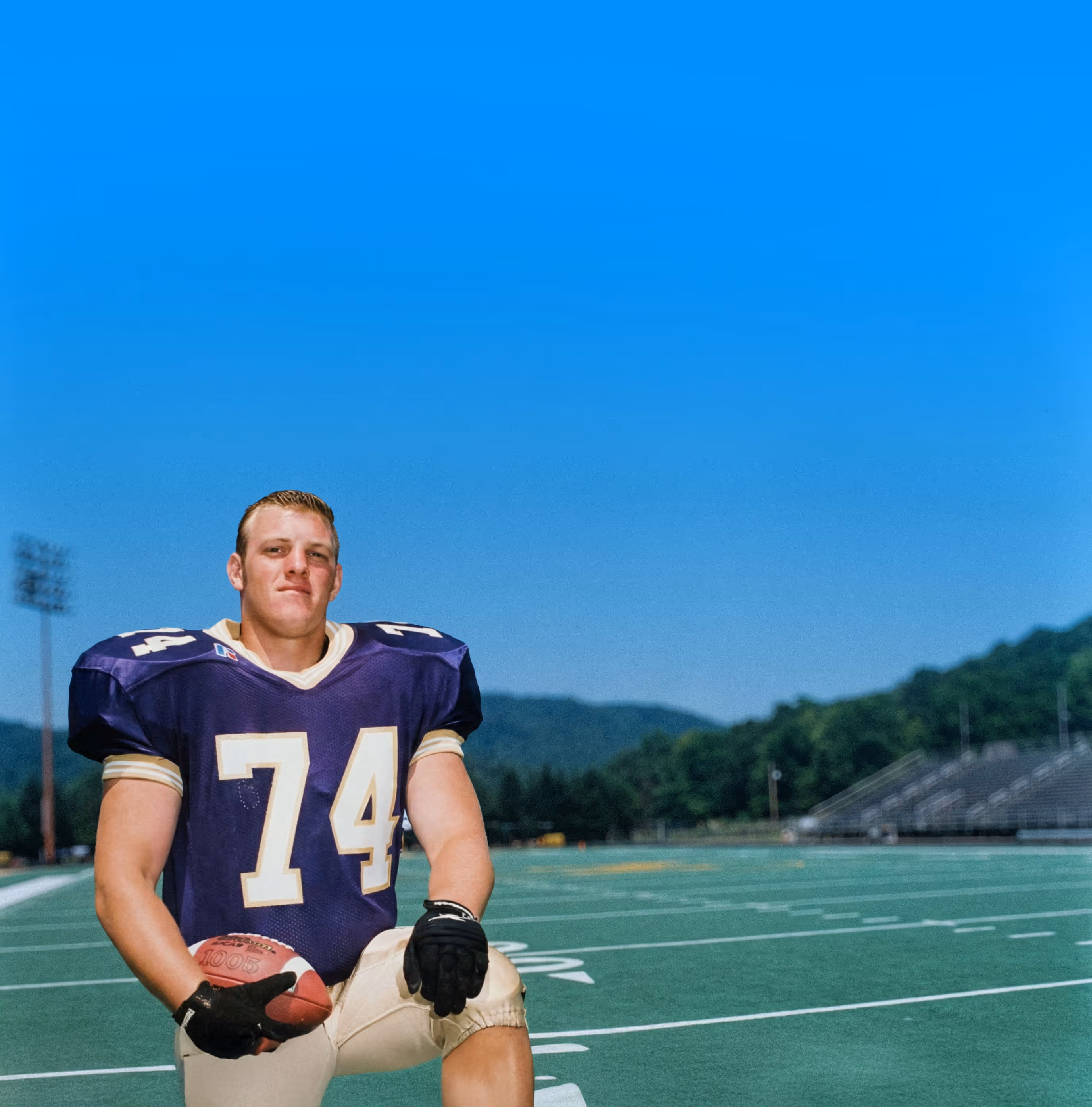 American football player, Paul Bondar, wearing a purple jersey numbered 74 kneeling on a football field holding a football.
