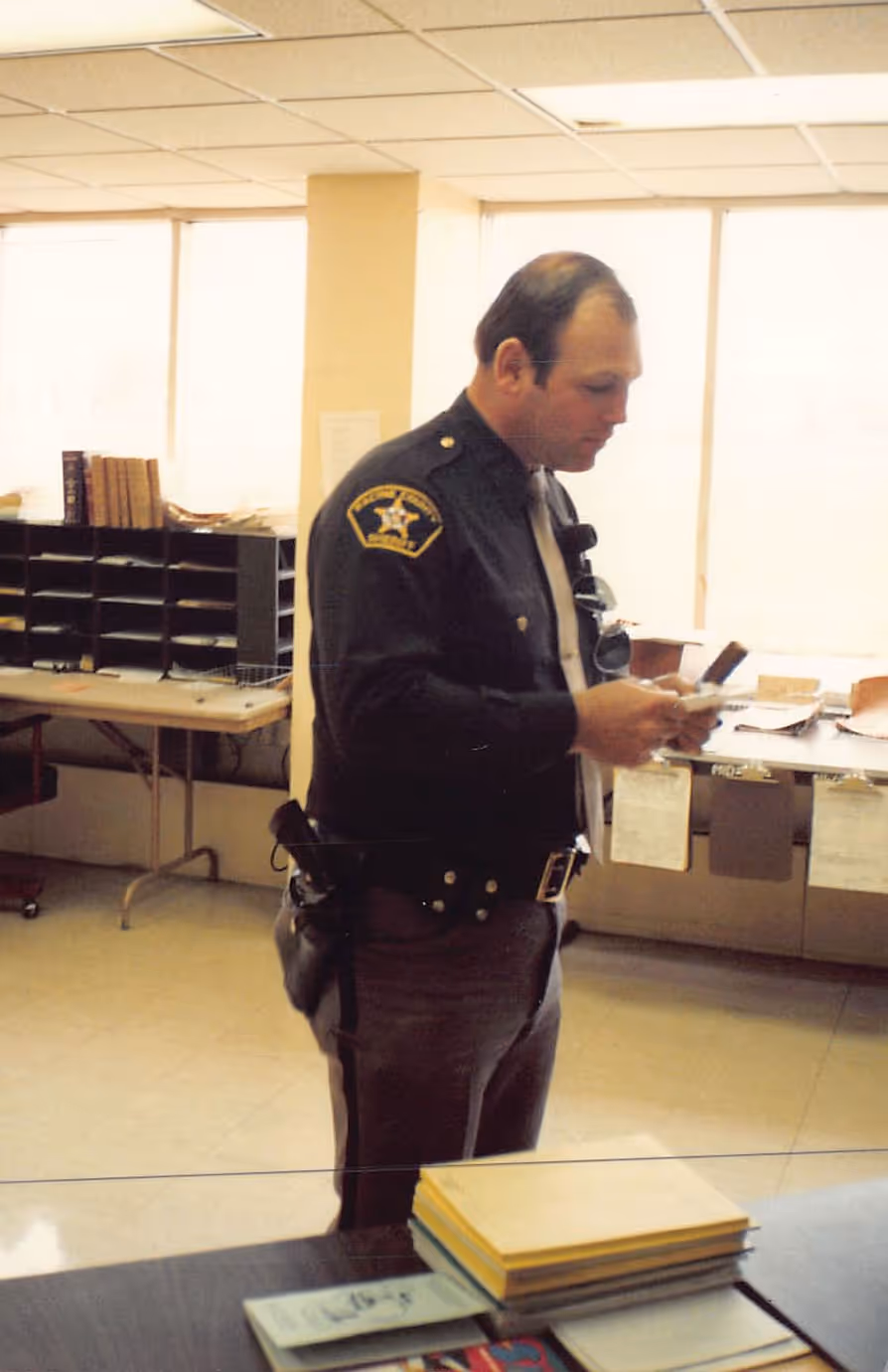 Police officer in uniform standing indoors reading a document with office furniture in the background.