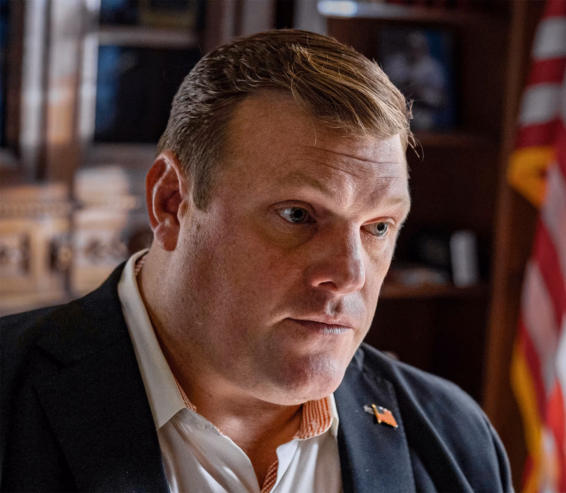 Close-up of Paul Bondar in a suit with a pin of the American flag on his lapel, looking thoughtfully to the side in an indoor setting with an American flag blurred in the background.