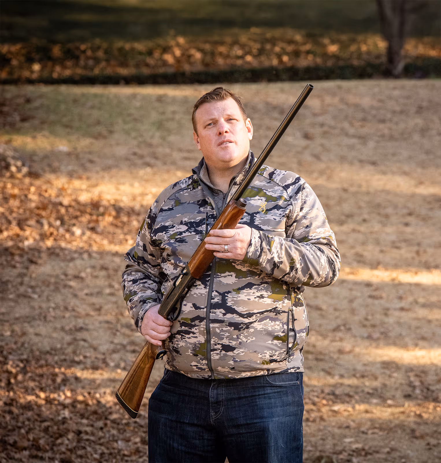 Paul Bondar wearing camouflage jacket holding a shotgun outdoors in a wooded area.